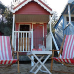 Beach hut with deck chairs and table set out in front. The beach hut is called "The Den" and is number 82 on Wells beach, Wells-next-the-Sea, North Norfolk. The hut is available to rent via Airbnb.