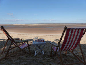 Deck chairs looking out from "The Den", Beach Hut 82, Wells-next-the-Sea, North Norfolk, over the wide expanse of sand of Wells beach at low tide, the channel in the middle distance - visible as a narrow strip - and the sea on the horizon.